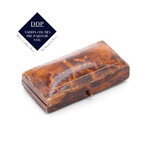 May include: A rectangular, antique leather box with a hinged lid. The box is a warm brown color with a mottled texture and a small brass clasp. The text "DDP Tariffs/Duties Pre-Paid For You" is in the upper left corner.