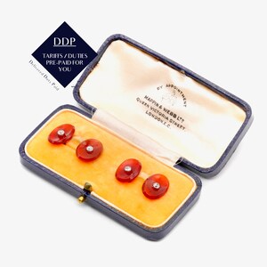 May include: A set of four oval-shaped cufflinks in a navy blue box. The cufflinks are a translucent reddish-orange color with a small, round, clear stone in the center. The box is open, revealing a yellow interior and text.