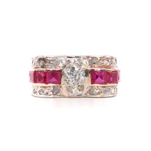 Puede incluir: Un anillo de banda ancha con un diamante central de talla brillante flanqueado por dos filas de rubíes rojos cuadrados, engastado en una banda de metal texturizada con numerosos diamantes pequeños en pavé en los bordes superior e inferior.