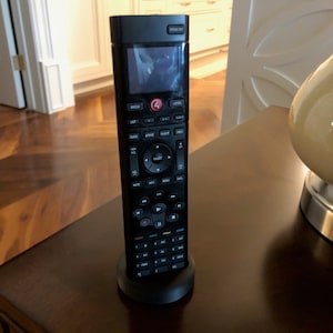 May include: A black universal remote control with a small screen and a variety of buttons, including volume, channel, and navigation controls. The remote is sitting on a black stand.