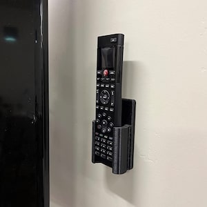 May include: A black remote control holder mounted on a white wall. The holder is made of 3D printed plastic and has a textured surface. The remote control is black and has a small screen and a variety of buttons.
