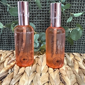 May include: Two glass spray bottles with copper-colored tops and labels. One is labeled "Lavender" and the other "Chamomile". The bottles are filled with a light orange liquid. The text "take well" is printed on the top of each bottle.
