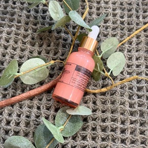 May include: A pink glass bottle with a gold lid and a dropper. The bottle is labeled "HYALURONIC ACID SERUM" and has a bee logo. The bottle is sitting on a brown textured surface.