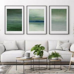 Puede incluir: Un salón moderno con un sofá blanco, una silla de madera y tres cuadros abstractos enmarcados de paisajes en tonos verdes y azules. La habitación está decorada con plantas en macetas y pequeñas mesas de centro redondas.