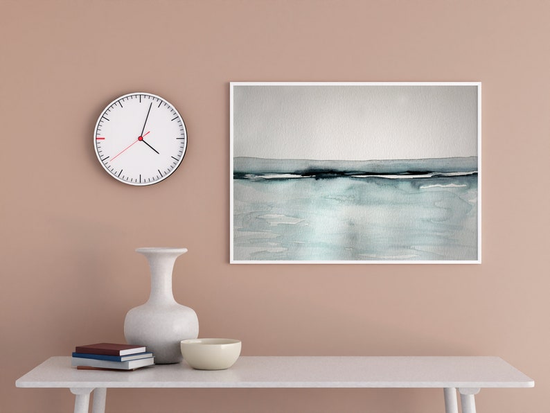 May include: A white clock with black hands and a red second hand hangs on a light brown wall above a white table. A white vase with a wide mouth sits on the table next to a white bowl. A framed watercolor painting of a blue and gray seascape hangs on the wall above the table.