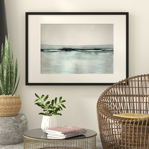 May include: A black framed watercolor painting of a seascape with a light blue and gray color scheme hangs on a white wall above a wicker chair and a glass-top coffee table with books.