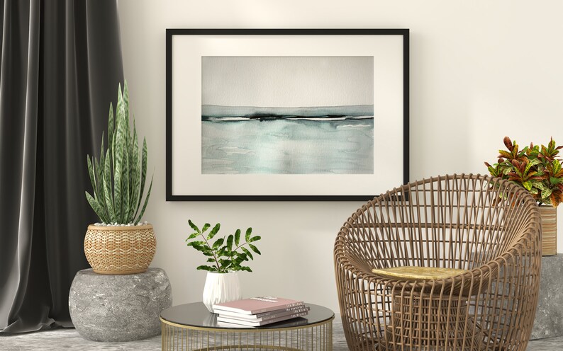 May include: A black framed watercolor painting of a seascape with a light blue and gray color scheme hangs on a white wall above a wicker chair and a glass-top coffee table with books.