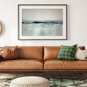 May include: A brown leather sofa with two pillows, one green and one white, sits in a living room with a framed abstract watercolor painting above it. A round wooden wall hanging and a potted plant are also visible.