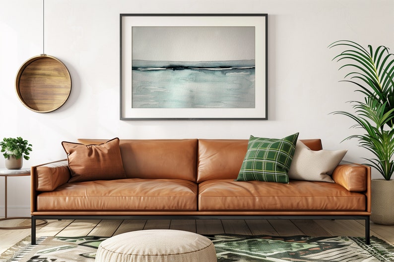 May include: A brown leather sofa with two pillows, one green and one white, sits in a living room with a framed abstract watercolor painting above it. A round wooden wall hanging and a potted plant are also visible.