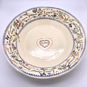 May include: A cream-colored ceramic bowl featuring a central heart design with the word Love. The rim is decorated with colorful floral patterns, butterflies, hummingbirds, and the text Love is patient, Love is kind, Love never fails.