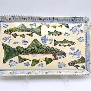 May include: Rectangular ceramic serving tray with a cream-colored surface, featuring painted green, red, and yellow fish. The tray has a blue border with wave and star patterns. Decorative blue fishing flies are also painted on the tray.