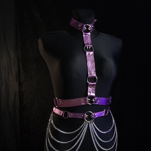 May include: A purple leather body harness with silver-tone metal hardware and chain accents. The harness includes a choker, shoulder straps, a waist belt, and dangling chains. The item is displayed on a black mannequin.