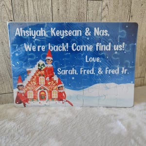 May include: A blue and white Christmas puzzle featuring three elves standing in front of a gingerbread house. The puzzle has the text "Ahsiyah, Keysean & Nas, We're back! Come find us! Love, Sarah, Fred, & Fred Jr."