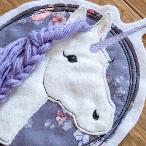 Pode incluir: Uma cabeça de unicórnio branca com uma crina e um chifre trançados roxos. O unicórnio é bordado em um fundo cinza com estampa floral. O desenho está em um suporte de feltro branco.