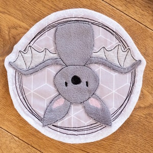 Può includere: Un pipistrello ricamato di colore grigio e bianco con orecchie rosa, appeso a testa in giù in un cerchio su uno sfondo di tessuto grigio e bianco.