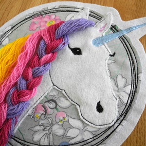 Peut inclure: Une tête de licorne blanche avec une crinière tressée arc-en-ciel et une corne bleue. La licorne est brodée sur un tissu gris à motif floral.