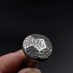 May include: Close-up of a silver metal cabinet knob with a hammered texture. The knob has a rounded, faceted design and is held against a dark background. The knob is approximately 1 inch in diameter.