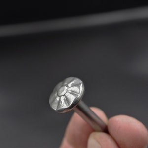 Peut inclure: Gros plan d'un clou argenté avec une tête décorative en forme de fleur. Le clou est tenu sur un fond gris foncé. La tête présente un motif texturé et détaillé. La tige du clou est lisse et cylindrique.