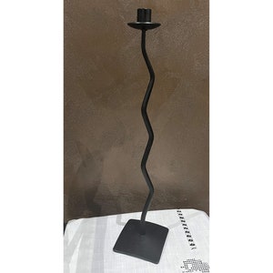 May include: A tall, black metal candlestick holder with a wavy stem and a square base. The top has a circular platform to hold a candle. The design is minimalist and modern, suitable for home decor.