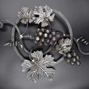 May include: A metal wall art piece featuring a grape vine design. The artwork includes a circular frame, grape clusters, and leaves. A corkscrew is also incorporated into the design. The metal has a silver finish.