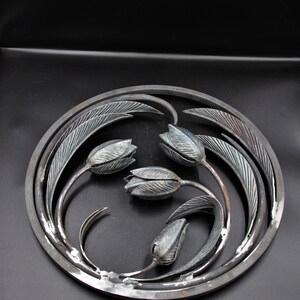 May include: A circular metal wall art piece with a design of four stylized tulips with leaves. The metal is a dark gray color with a slightly textured finish.