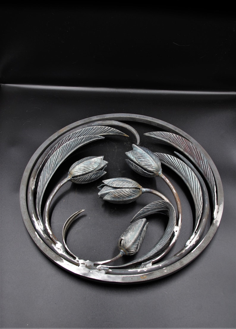 May include: A circular metal wall art piece with a design of four stylized tulips with leaves. The metal is a dark gray color with a slightly textured finish.