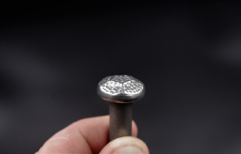 May include: Close-up of a silver nail with a hammered, faceted head. The nail's head has a textured surface and a rounded shape. The nail is held against a dark background, highlighting its metallic sheen.