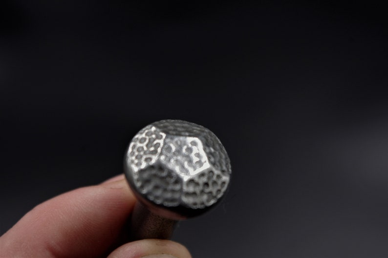 May include: Close-up of a silver metal cabinet knob with a hammered texture. The knob has a rounded, faceted design and is held against a dark background. The knob is approximately 1 inch in diameter.