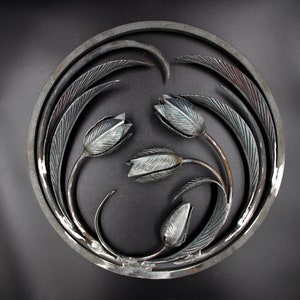 May include: A circular metal wall art piece with a design of stylized tulips and leaves. The piece is made of iron and has a dark gray finish.
