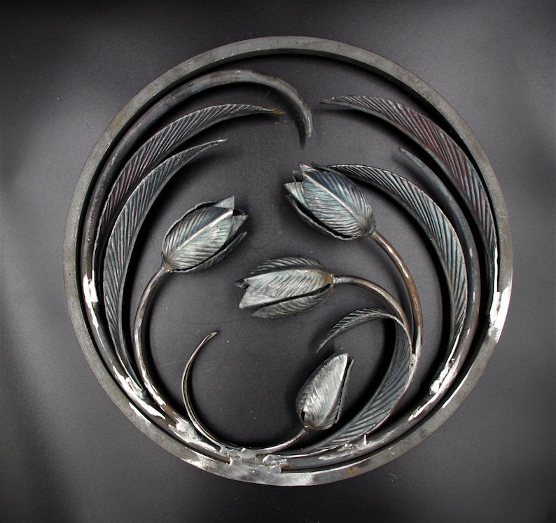 May include: A circular metal wall art piece with a design of stylized tulips and leaves. The piece is made of iron and has a dark gray finish.