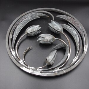 May include: A circular metal wall art piece featuring four stylized tulips with leaves. The tulips are made of metal and have a textured finish. The piece is a dark gray color.
