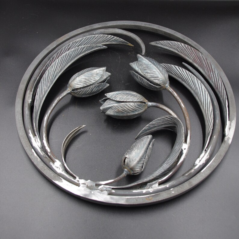 May include: A circular metal wall art piece featuring four stylized tulips with leaves. The tulips are made of metal and have a textured finish. The piece is a dark gray color.