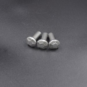 May include: Three silver-colored rivets with textured, rounded heads. The rivets are arranged in a cluster on a dark gray surface. The heads have a hammered or dimpled appearance, suggesting a decorative or industrial design.