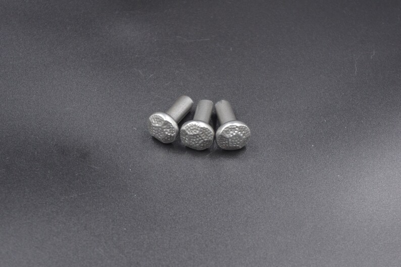 May include: Three silver-colored rivets with textured, rounded heads. The rivets are arranged in a cluster on a dark gray surface. The heads have a hammered or dimpled appearance, suggesting a decorative or industrial design.