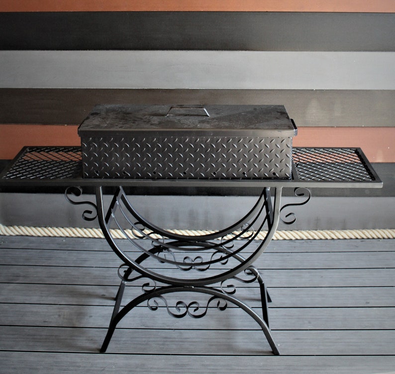 May include: Black metal grill with a diamond plate lid and a decorative stand. The grill is sitting on a wooden deck.