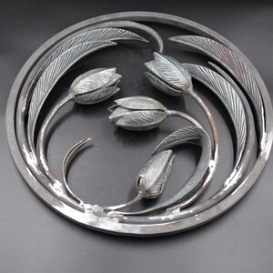 May include: A circular metal wall art piece with a design of four stylized tulips with leaves. The piece is made of metal and has a silver finish.