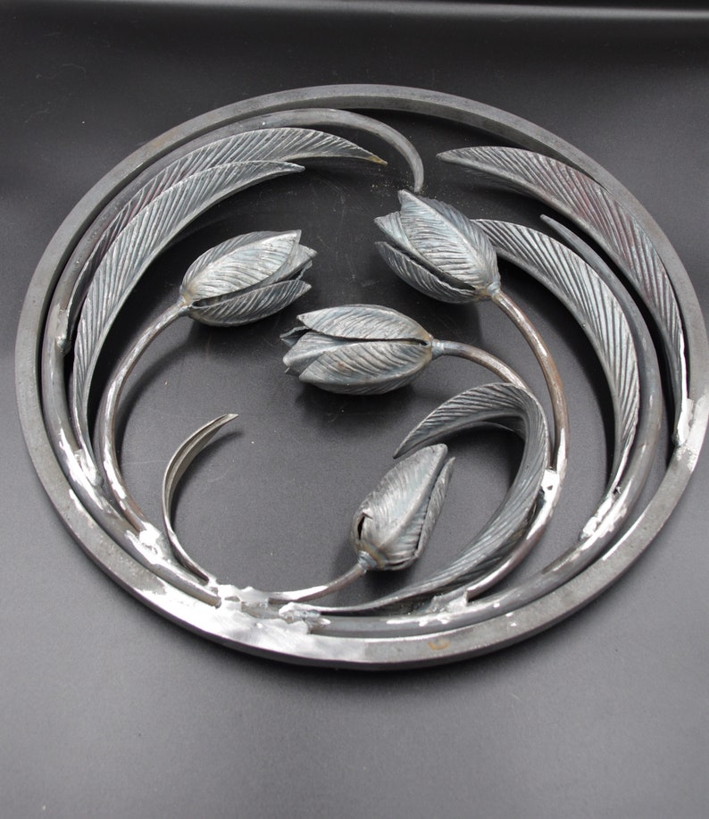 May include: A circular metal wall art piece with a design of four stylized tulips with leaves. The piece is made of metal and has a silver finish.