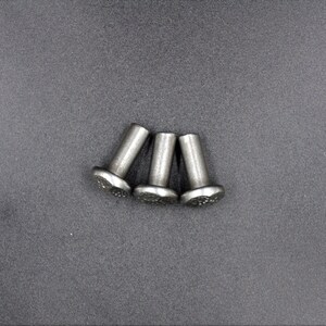 May include: Three silver-colored rivets with a textured, flower-like head. The rivets are cylindrical with a slightly wider head. They are arranged on a gray surface.