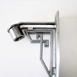 Könnte beinhalten: Eine silberfarbene Metallhalterung mit Art-Deco-Design. Die Halterung hat ein zylindrisches Element und ein dekoratives geometrisches Muster. Sie ist auf einer flachen, rechteckigen Basis montiert.