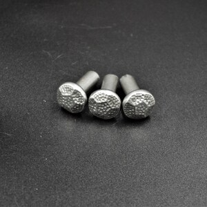 May include: Three silver-colored decorative nails with textured, rounded heads. The nails are arranged in a slight arc against a dark background. The heads have a hammered appearance, adding a rustic touch to the hardware.