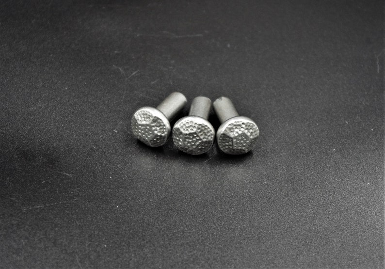 May include: Three silver-colored decorative nails with textured, rounded heads. The nails are arranged in a slight arc against a dark background. The heads have a hammered appearance, adding a rustic touch to the hardware.