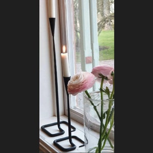 Könnte beinhalten: Eine Fensterdekoration mit zwei schwarzen Metallkerzenhaltern mit brennenden weißen Kerzen. Eine klare Glasvase enthält rosa und weiße Blumen. Die Szene spielt sich vor einem Fenster mit Blick ins Grüne ab.