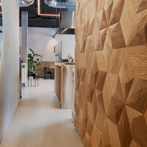 May include: Interior view of a modern space with a geometric wooden wall. The hallway features a light-colored floor and walls, with a large, oval-shaped mirror on the left. Overhead, exposed ductwork and lighting fixtures add an industrial touch.