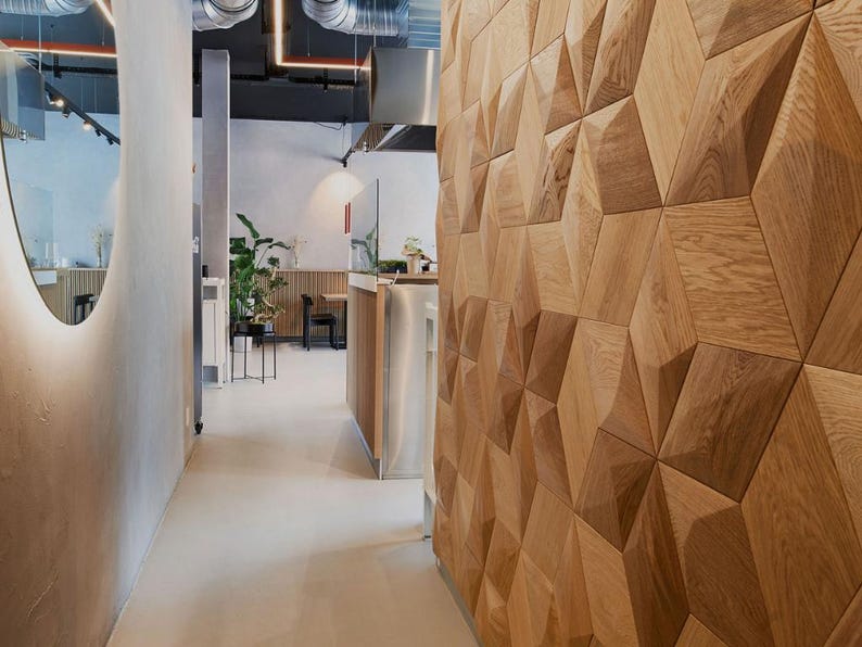 May include: Interior view of a modern space with a geometric wooden wall. The hallway features a light-colored floor and walls, with a large, oval-shaped mirror on the left. Overhead, exposed ductwork and lighting fixtures add an industrial touch.