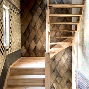 May include: Wooden staircase with light-colored steps and a matching handrail. The wall features a geometric pattern of diamond-shaped wooden panels in varying shades of brown. The adjacent wall is covered in light-colored tiles, and a small window is visible.