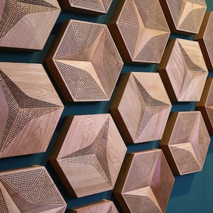 Op de afbeelding: Een muur bedekt met zeshoekige houten akoestische panelen. De panelen zijn lichtbruin en hebben een geometrisch patroon van kleine gaatjes.