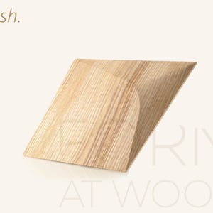 Op de afbeelding: Een lichtgekleurd essenhouten object met een geometrische, hoekige vorm, voorzien van een plat bovenoppervlak en een gebogen zijprofiel. Het woord Ash staat in de linkerbovenhoek en de tekst ERMI AT WOOD is zichtbaar in de rechterbenedenhoek.