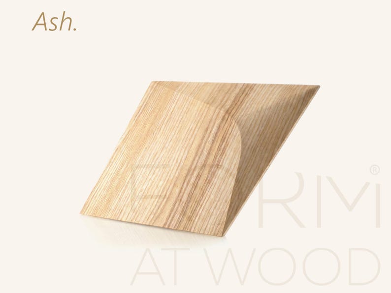 Op de afbeelding: Een lichtgekleurd essenhouten object met een geometrische, hoekige vorm, voorzien van een plat bovenoppervlak en een gebogen zijprofiel. Het woord Ash staat in de linkerbovenhoek en de tekst ERMI AT WOOD is zichtbaar in de rechterbenedenhoek.