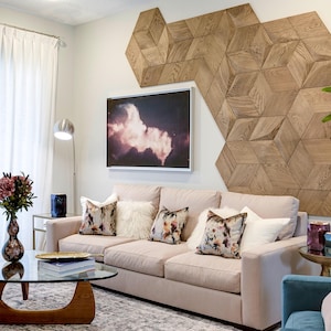 Op de afbeelding: Een beige bank met bloemmotief en witte imitatiebont kussens, geplaatst voor een muur met een grote, geometrische houten paneelinstallatie bestaande uit zeshoekige en ruitvormige tegels met een zichtbare houtnerfstructuur.