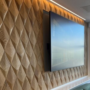 Textured accent wall in a modern interior, featuring 3D wall panels in a diamond pattern made of natural wood. The wall decor surrounds a large TV screen, combining wood wall art with functional design. This wood wall paneling enhances the room’s ambiance with stylish LED lights above, creating a warm and inviting atmosphere. Ideal for living room decor or office wall paneling.